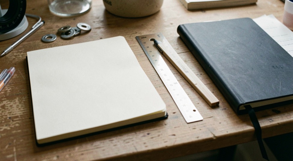 A wooden desk holds a blank notebook, drafting ruler, metal washers, and a closed folder in soft light.